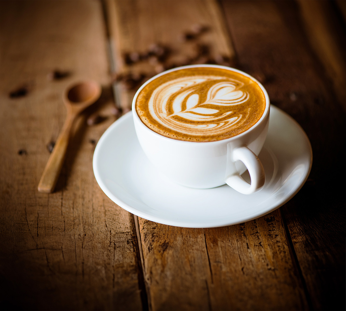 A cup of Cappiccino on a wooden table, around the cup are coffee beans and a wooden spoon