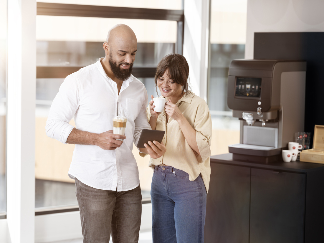 Zwei Personen stehen an einer Kaffeemaschine und betrachten gemeinsam ein Tablet.